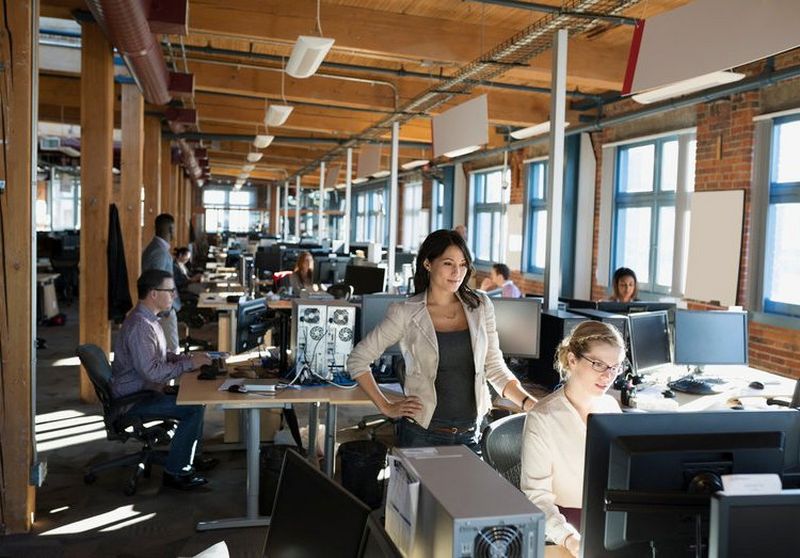 Woman with hand on her hip stands over another woman working at a large computer screen in an office warehouse setting.